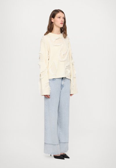 White sweater with textured, frayed detailing. Paired with high-waisted light blue wide-leg jeans and pointed black heels.