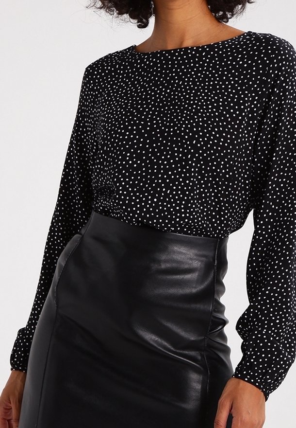 Femme portant un chemisier noir à manches longues à pois blancs, rentré dans une jupe en cuir noire taille haute.