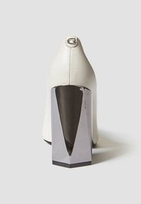 Chaussure à talon haut blanche avec une tige en cuir texturé et un talon métallique angulaire et brillant. Présente un logo argenté circulaire à l'arrière.