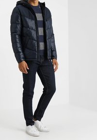 Marinblå vadderad jacka med dragkedja framtill och huva, med horisontell sömnad. Kombinerad med mörka jeans och vita sneakers.