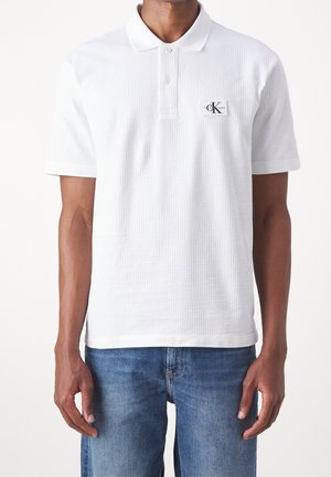 Mężczyzna noszący białą, teksturowaną koszulkę polo z krótkim rękawem, z małą naszywką z logo CK Jeans na piersi oraz niebieskie dżinsy.