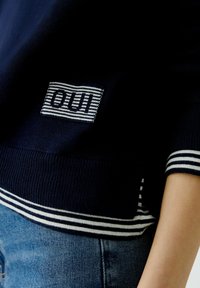 Marineblauer Pullover mit gestreiften weißen Akzenten am Saum und an den Ärmeln, versehen mit einem kleinen rechteckigen Logopatch. Weiches, strukturiertes Strickmaterial.