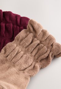 Twee gebreide beenwarmers: één in bordeauxrood met een geribbelde textuur en één in beige met golvende, gerimpelde randen, beide zacht en elastisch.