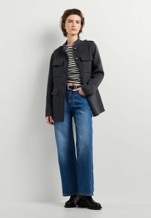 Jong persoon draagt een donkergrijze jas in militaire stijl, gestreept cropped topje, hoog getailleerde blauwe jeans en zwarte enkellaarzen, staand met één hand in de zak.