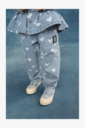 Pantalon en denim bleu clair avec motifs de cœurs blancs, doté d'une étiquette noire de marque. Associé à des baskets rayées bleu et blanc sur un fond bleu.