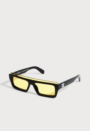 Lunettes de soleil rectangulaires noires avec des verres jaunes et une fine barre dorée sur le dessus, avec un petit logo blanc sur la branche.