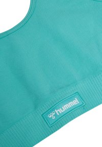Nahtloser türkisfarbener Sport-BH mit gerippter Struktur und einem "hummel" Logo-Patch, das nahe am unteren Bund angenäht ist.