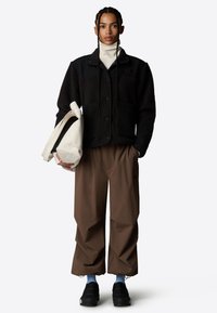 Chaqueta de forro polar negra con un acabado texturizado, cuello alto crema, pantalones anchos marrones y zapatos negros sin cordones; sosteniendo un bolso crema.