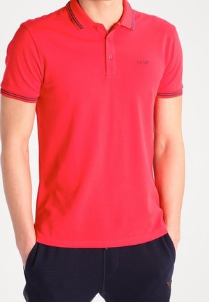 Punainen poolopaita, jossa on kaksi nappia sisältävä aukko, resoroitu kaulus ja kontrastivärisiä raitoja. Vasemmassa rinnassa logo. Silkkinen kangas.