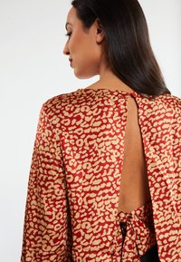 Rood en beige gestippelde blouse met een lage open rug, voorzien van een striksluiting en lange mouwen. Soepele stof met een metallic glans.