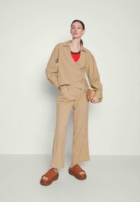 Una donna si erge indossando un top avvolto beige e pantaloni, una camicia rossa a strati, sandali con zeppa marroni e tiene in mano una piccola borsa a tracolla intrecciata.
