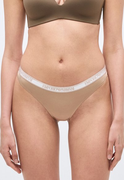 Γυναίκα που φορεί μπεζ εσώρουχο χαμηλής μέσης και ταίριαστο σουτιέν με λευκή ελαστική ζώνη μέσης με το λογότυπο "Emporio Armani" σε απλό φόντο.