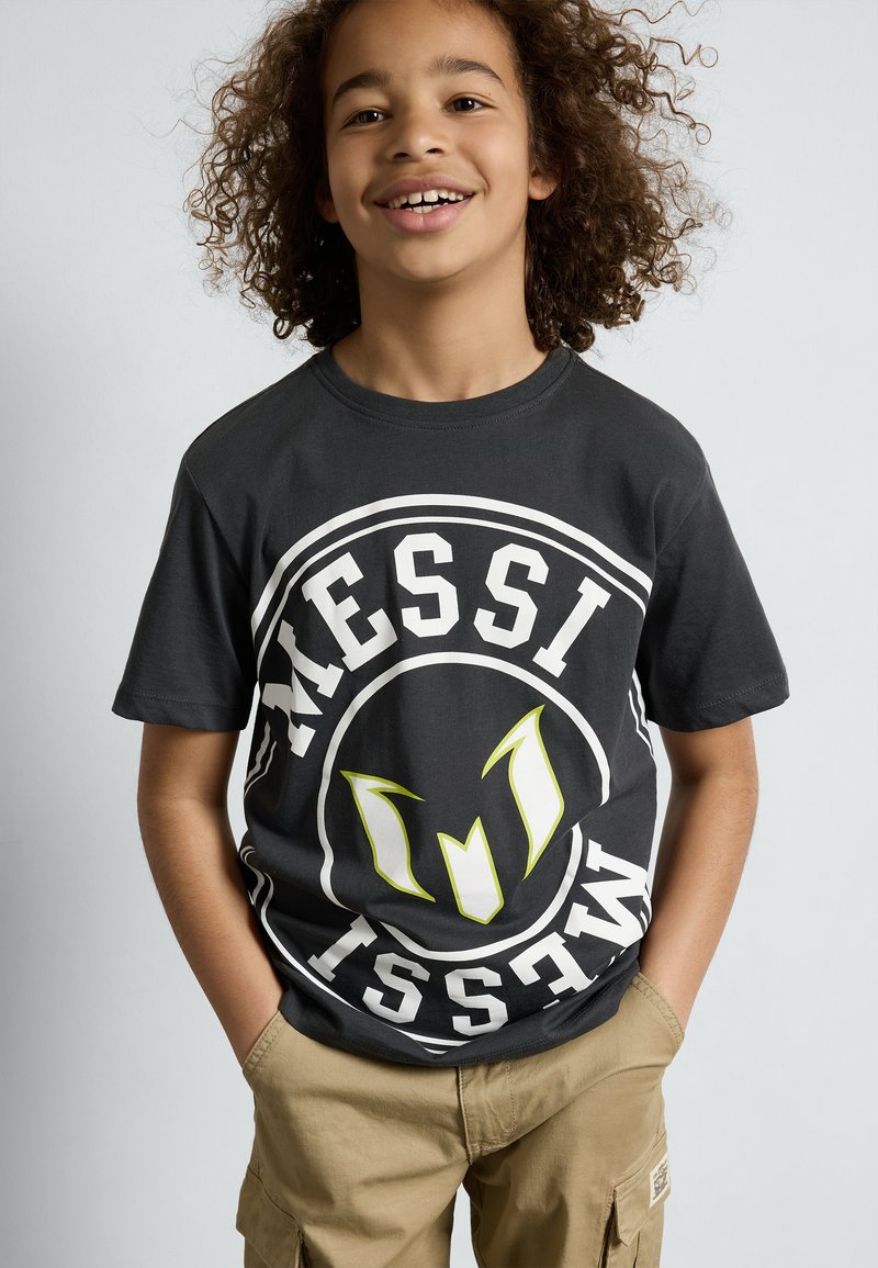Svart bomull t-skjorte med et stort, sirkulært logo i hvitt med grønne aksenter og teksten "MESSI." Kombinert med beige cargo-shorts.