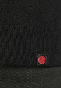Tissu tricoté noir avec une petite étiquette noire affichant un cercle rouge cousu près du bord inférieur.