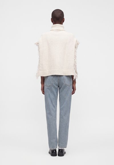 Personne portant un pull en tricot sans manches crème avec des franges, un jean bleu clair et des chaussures noires, debout de dos sur fond blanc.