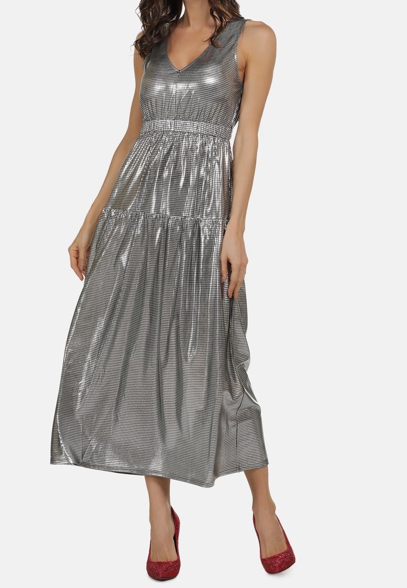 Femme portant une robe midi métallique argentée sans manches avec une taille froncée, associée à des talons hauts rouges pailletés, debout sur un fond blanc.