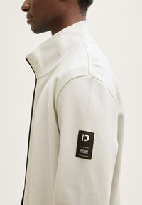 Veste gris clair avec un col haut et une fermeture éclair complète. Dotée d'un tissu texturé et d'une étiquette noire sur la manche gauche avec un texte blanc.