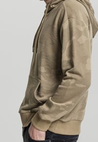 Mikina s kapucí v béžové barvě s jemným maskáčovým vzorem. Obsahuje klokánkovou kapsu a nastavitelné šňůrky. Materiál je smíchaný bavlněný.