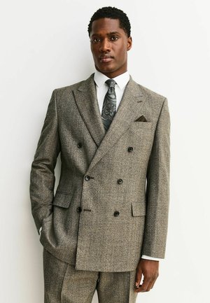 Man wearing a gray double-breasted suit with a white shirt and patterned tie, standing against a plain light background.