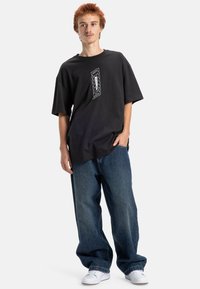Černé oversized tričko s bílým grafickým textem, spárované s volnými modrými džíny a bílými teniskami.