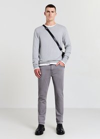 Graues Sweatshirt mit Rundhalsausschnitt, getragen über einer weißen Schicht, kombiniert mit hellgrauen Hosen und schwarzen Ankle Boots. Schwarz Schultergurt enthalten.