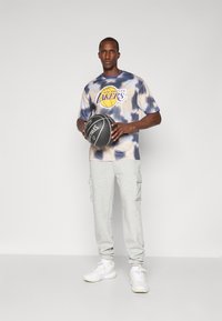 Grafisk T-shirt med blå og creme tie-dye mønster og "Los Angeles Lakers" logo, parret med grå cargo bukser og hvide sneakers. Holder en basketball.