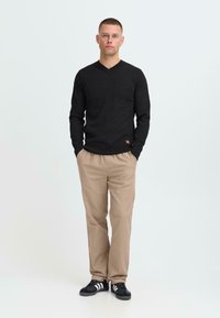 Man met kort haar staat met zijn handen in de beige broekzakken, gekleed in een zwart V-hals sweater met lange mouwen en zwarte sneakers met witte strepen.