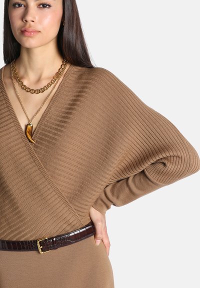 Femme portant un pull cache-cœur marron côtelé avec de larges manches, une ceinture marron foncé et deux colliers en or, dont un avec un pendentif en forme de dent.