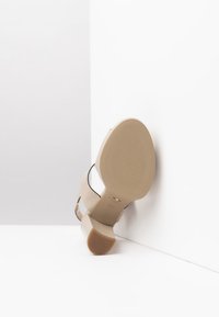 Sandale à talon beige avec un dessus lisse, des accents noirs et une semelle en caoutchouc texturée. Le design comprend des côtés ouverts et un bout arrondi.