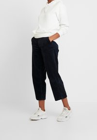 Person stående i vit hoodie, svarta korta kordbyxor och vita sportskor med händerna i fickorna.