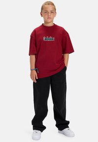 Bordeaux oversized T-shirt met geborduurde tekst in groen en blauw. Gecombineerd met zwarte brede broek en witte sneakers.