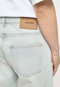 Jeans in denim blu chiaro con una toppa in pelle marrone etichettata "EIGHTYFIVE", con una sola tasca posteriore e dettagli di cucitura visibili.