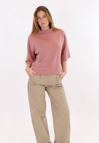 Giovane donna che indossa un top largo color malva con maniche a tre quarti e pantaloni beige a gamba larga, in piedi con le mani nelle tasche su sfondo bianco.