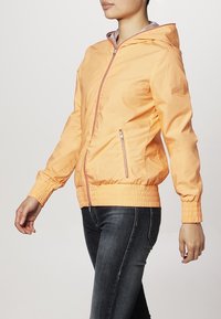 Giacca arancione con zip e cappuccio, polsini e fascia elastici; presenta due tasche laterali con zip eleganti e una superficie liscia e lucida.