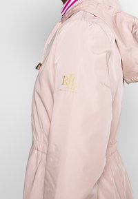 Ljusrosa, huvan jacka med elastiska ärmslut och rynkning i midjan. Har en guldlogotyp på vänster ärm och en randig krage.