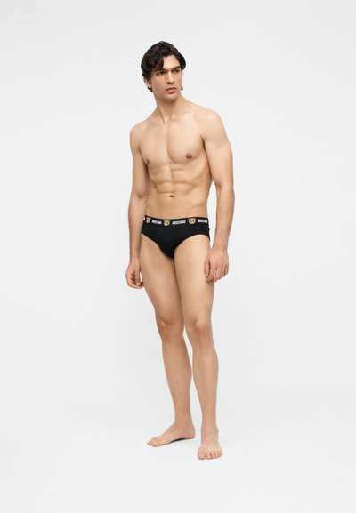 Άνδρας χωρίς πουκάμισο, ξυπόλητος, φοράει μαύρα εσώρουχα με πρόσωπα αρκουδάκια και το λογότυπο «Moschino» στη μέση, σε απλό φόντο.