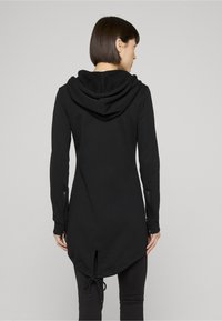 Fekete cipzáras kapucnis pulóver hátul hasítékkal, hosszú ujjakkal és zsinóros szegéllyel. Puha tapintású anyagból készült, tágas kapucnival a kényelemért.