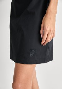 Musta DKNY-mekko, jossa on tyylikäs muotoilu ja brodeerattu logo helman kohdalla. Tasainen pinta ja polven pituinen leikkaus näkyvissä.