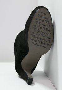 Botín de ante negro de tacón alto con puntera afilada y diseño de tobillo curvado. La suela presenta el texto de la marca en relieve y el tamaño número 36.