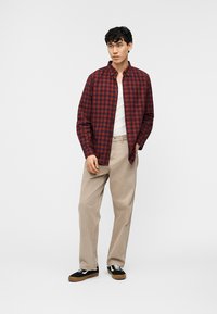 Νέος άνδρας όρθιος, φοράει κόκκινο και μαύρο καρό πουκάμισο πάνω από λευκή φανέλα, μπεζ παντελόνι και μαύρα αθλητικά παπούτσια με λευκά κορδόνια.