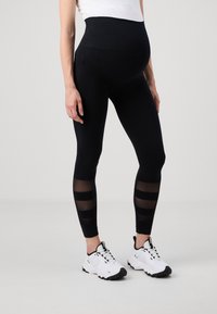 Leggings de maternité noirs avec une taille haute, ornés de panneaux rayés transparents sur le bas des jambes. Portés avec des baskets de sport blanches.