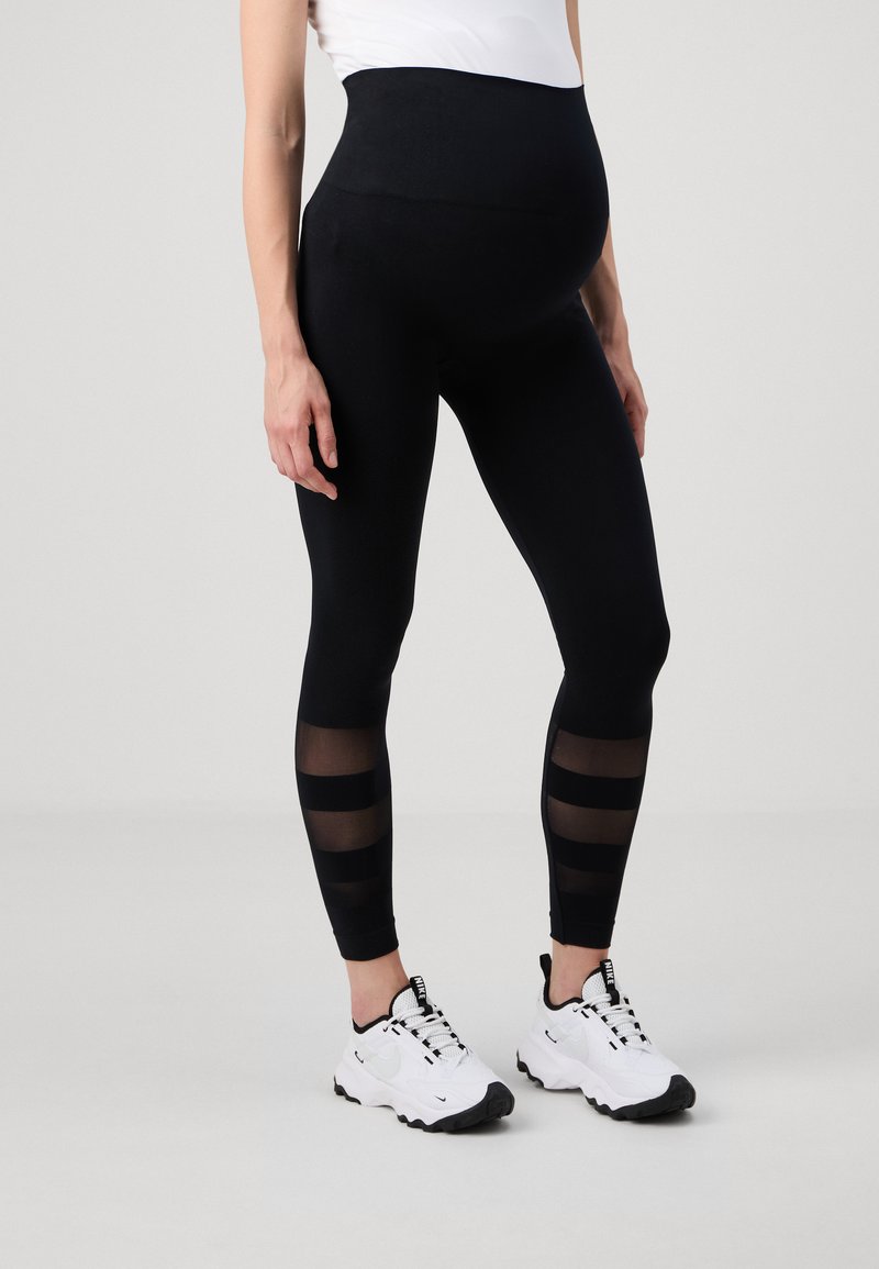 Leggings de maternité noirs avec une taille haute, ornés de panneaux rayés transparents sur le bas des jambes. Portés avec des baskets de sport blanches.