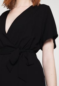 Vestido negro cruzado con escote en V pronunciado, mangas cortas y detalle de cintura atada. Tejido suave con una textura sutil.