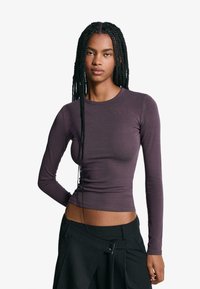 Long-sleeve, fitted top in dark purple, made of soft fabric. Features a round neckline and smooth texture, paired with high-waisted black pants.