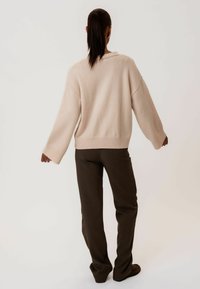 Jasnoróżowy, oversize sweter z opadającymi ramionami, ribbowym wykończeniem i szerokimi rękawami, zestawiony z ciemnobrązowymi, luźnymi spodniami.