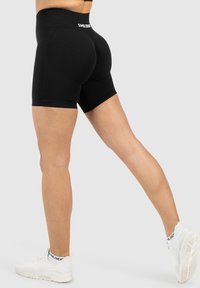 Model in zwarte shorts en witte sneakers, staand in profiel tegen een grijze achtergrond, die de pasvorm en stijl van de kleding toont.
