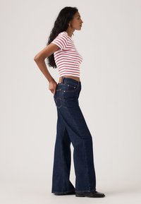 Pika tumejuustega naine, kes kannab punase-valgete triipudega lühikeste varrukatega topi ja tumedat sinist laiade säärtega teksapükse, seisab ja tõmbab pükste taskut.