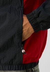 Veste coupe-vent noire et rouge avec une finition texturée, dotée de poignets élastiques, d'une poche zippée avec un tirette personnalisé et d'une poche latérale pour les mains.