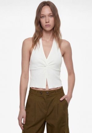 Młoda kobieta z prostymi włosami do ramion, ubrana w biały bezrękawnik z dekoltem w serek i oliwkowe spodnie z wysokim stanem.