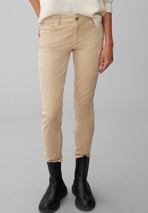 Schmal geschnittene beige Hose kombiniert mit schwarzen Stiefeletten, getragen mit einem weißen Strickpullover, gezeigt von der Hüfte bis zu den Füßen vor einem grauen Hintergrund.